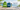 Three personalized gift items displayed outdoors on a wooden deck with a scenic ocean backdrop: a green drawstring bag, a blue tote bag, and a clear water bottle, all printed with the name GEORGE and promotional text on the right.